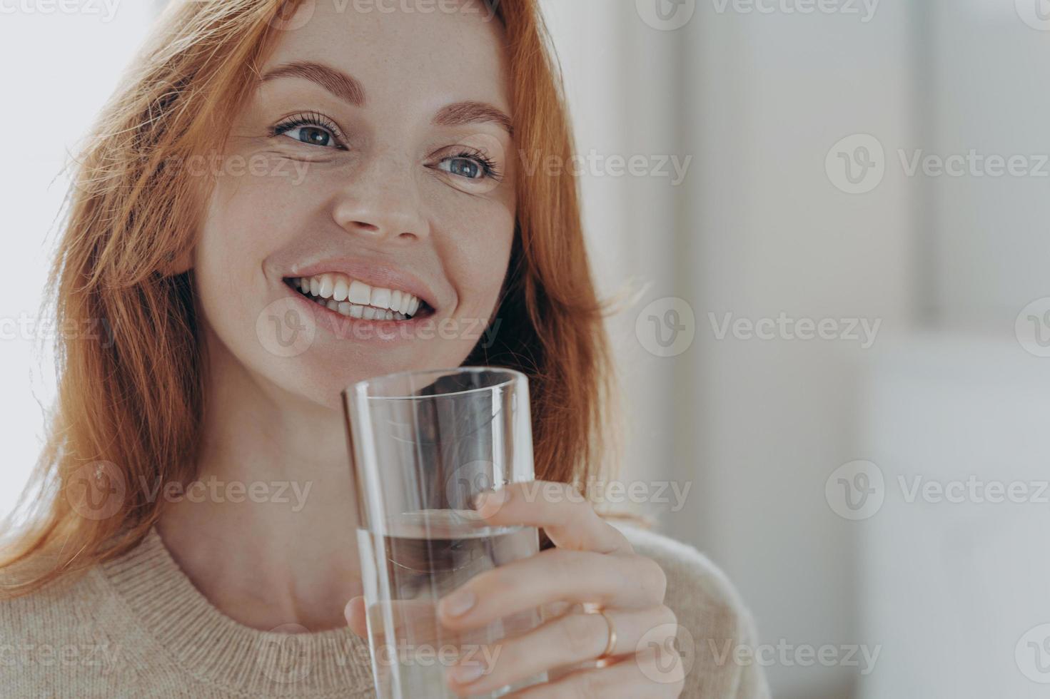 retrato de una feliz y saludable mujer pelirroja sosteniendo un vaso transparente de agua mineral pura foto