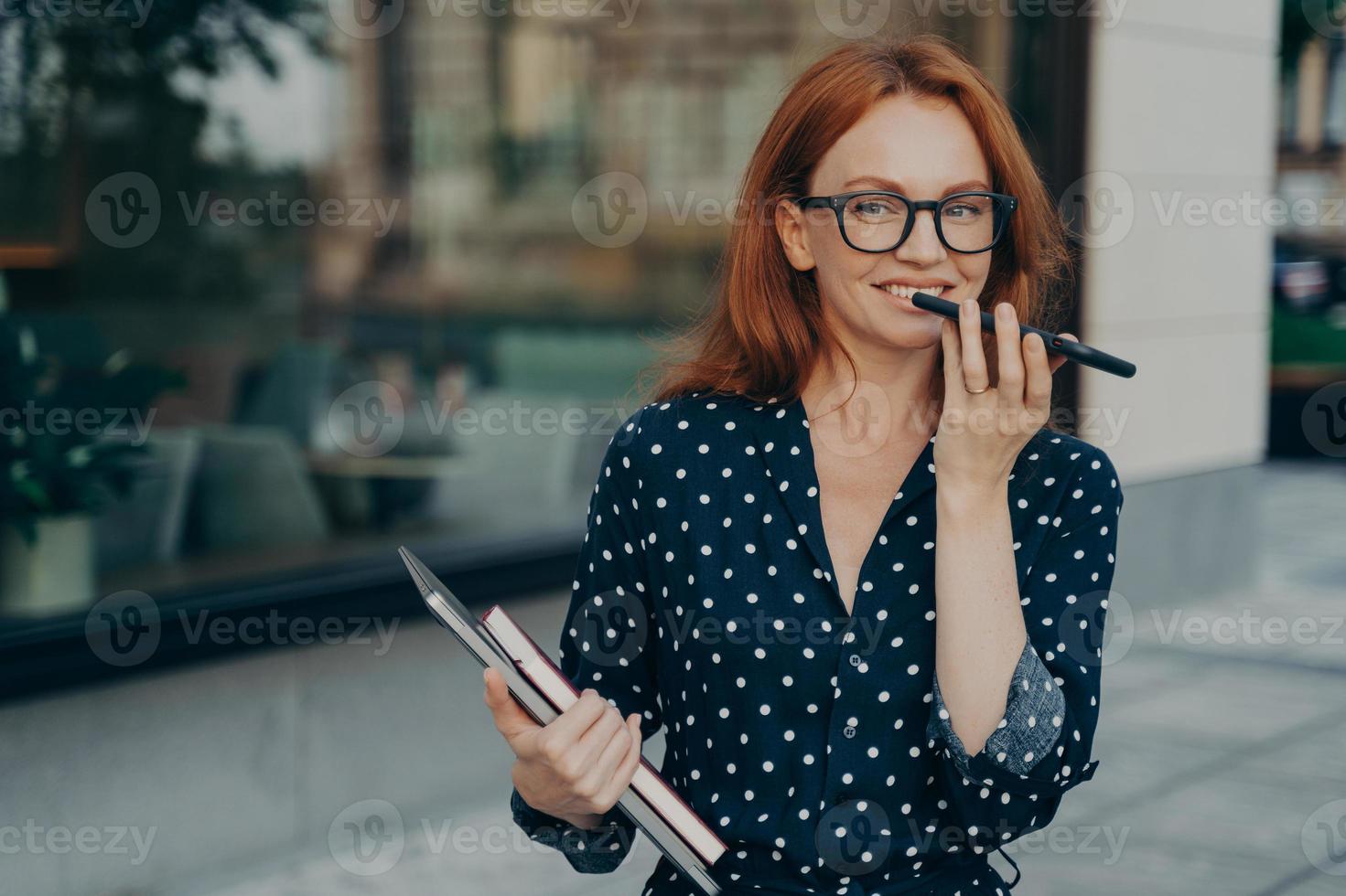 Una exitosa mujer de negocios pelirroja graba un mensaje de voz y mantiene el teléfono inteligente cerca de la boca foto