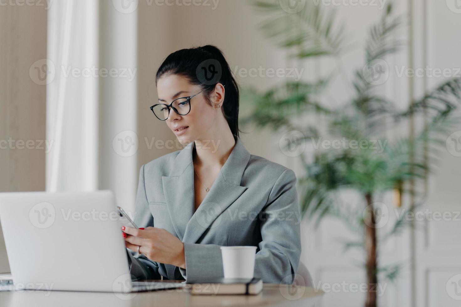Gorgeous busineswoman types message on smartphone, works on new publication, poses in front of laptop computer, involved in working process, sits at desktop, drinks aromatic coffee, uses internet photo