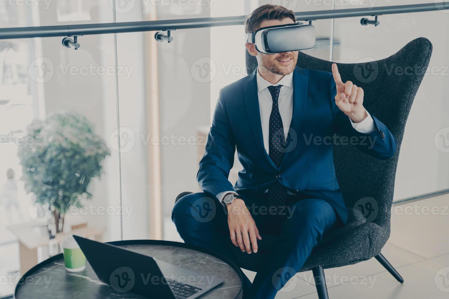 Smiling businessman using VR headset glasses while sitting in office lobby and working on laptop photo
