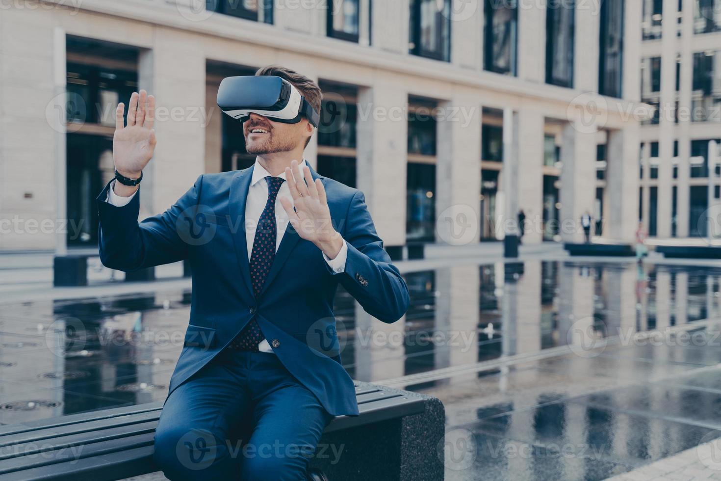 Man in VR goggles trying to reach something in virtual reality with his hands while sitting outdoors photo