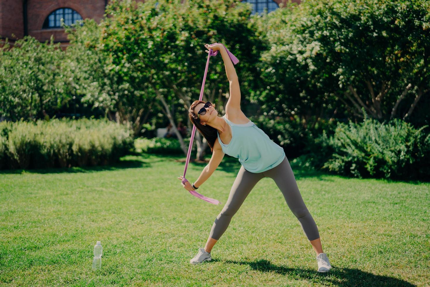 Full length shot of active woman stretches arms with fitness gum has good flexibility wears t shirt leggings and sneakers has workout with resistance band poses outdoor on green lawn during summer photo