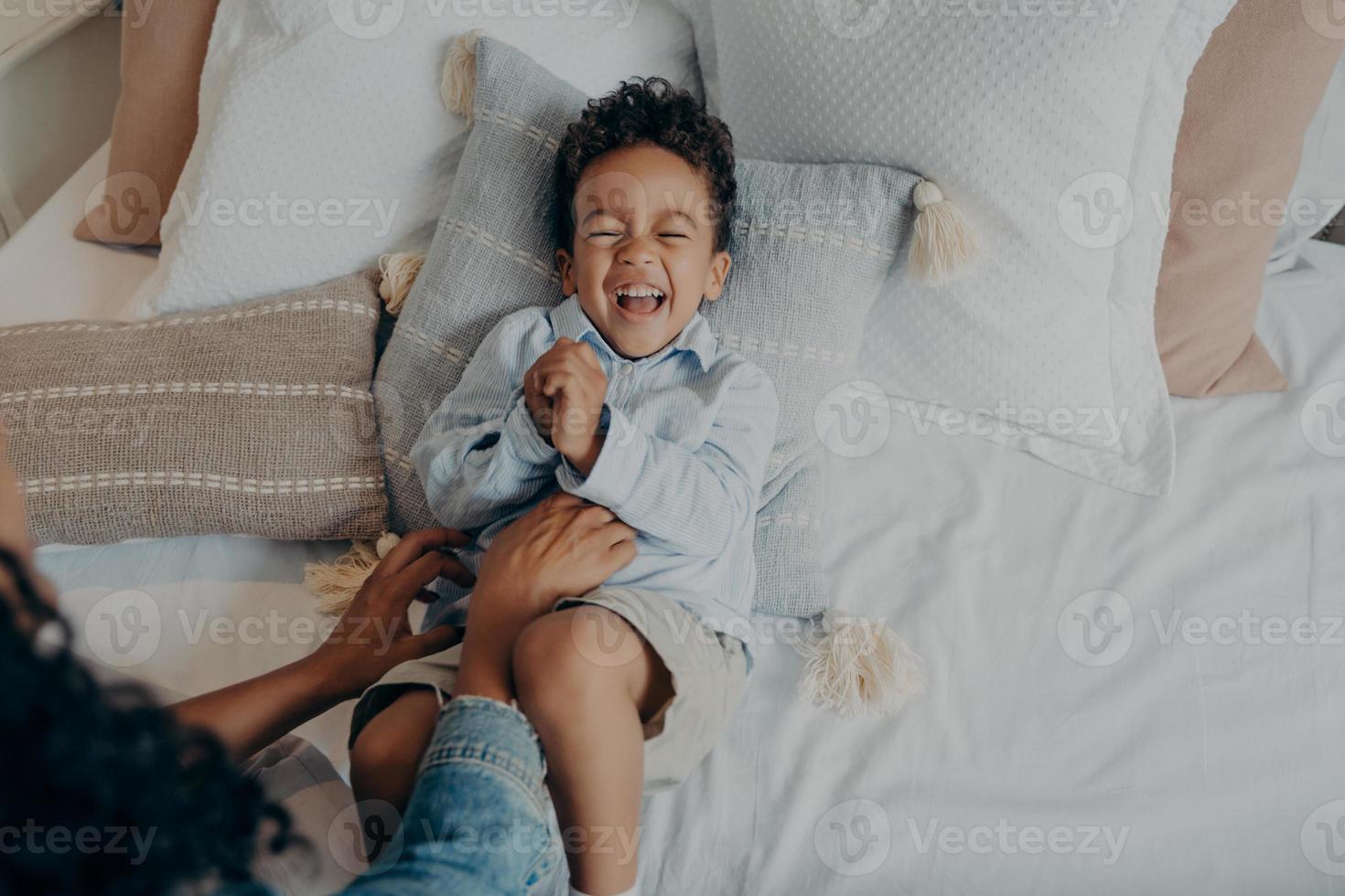 Adorable little son lying on pillows in bed and laughing while mom tickling him photo