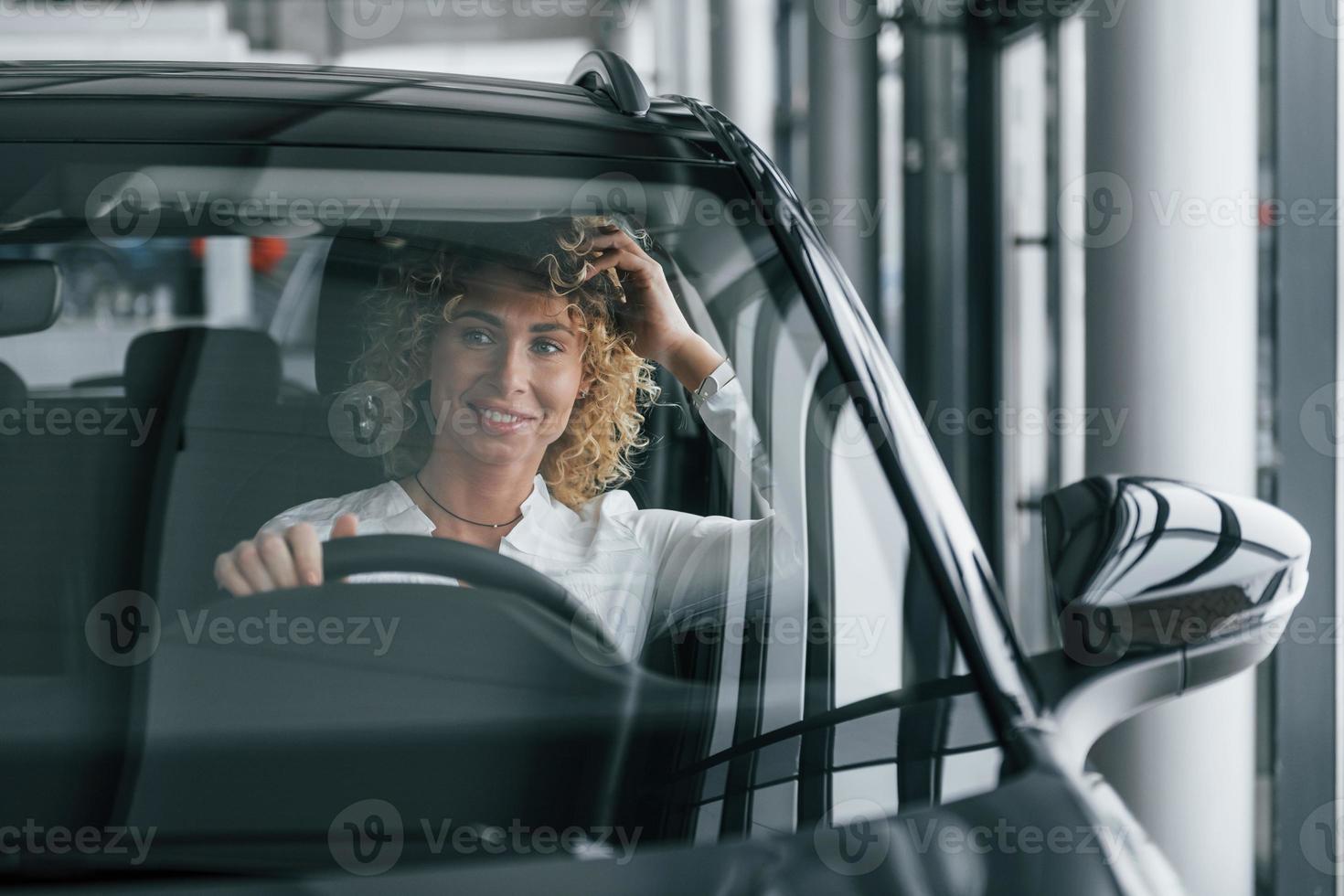 Sitting at driver's seat. Woman with curly blonde hair is in autosalon photo
