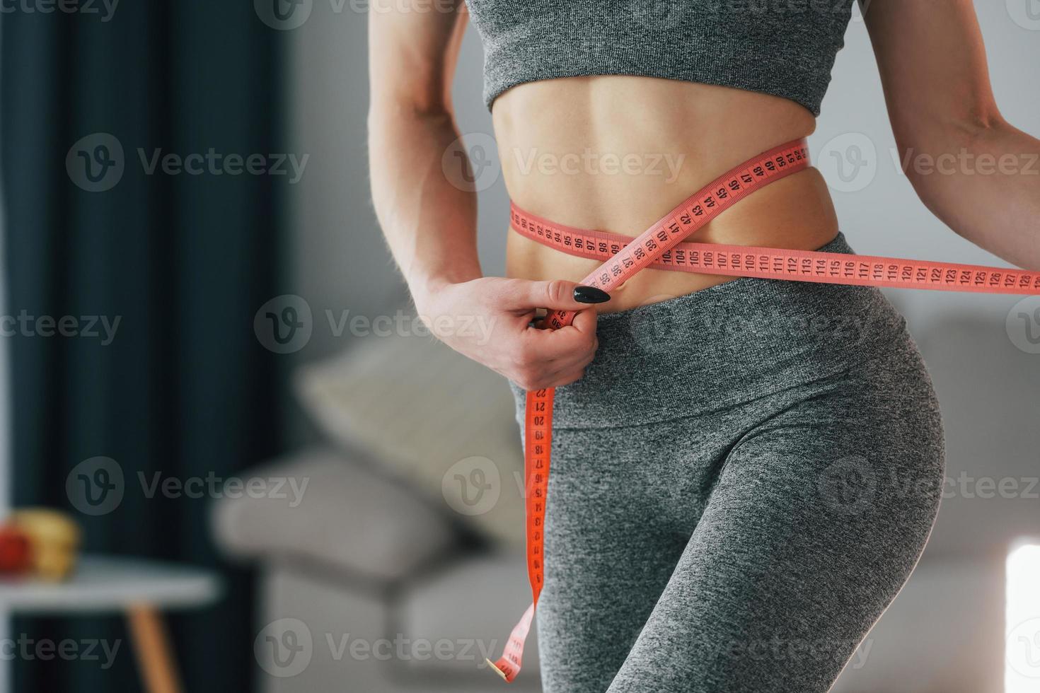 Pink colored measuring tape around the waist. Young woman with slim body type and in yoga clothes is at home photo