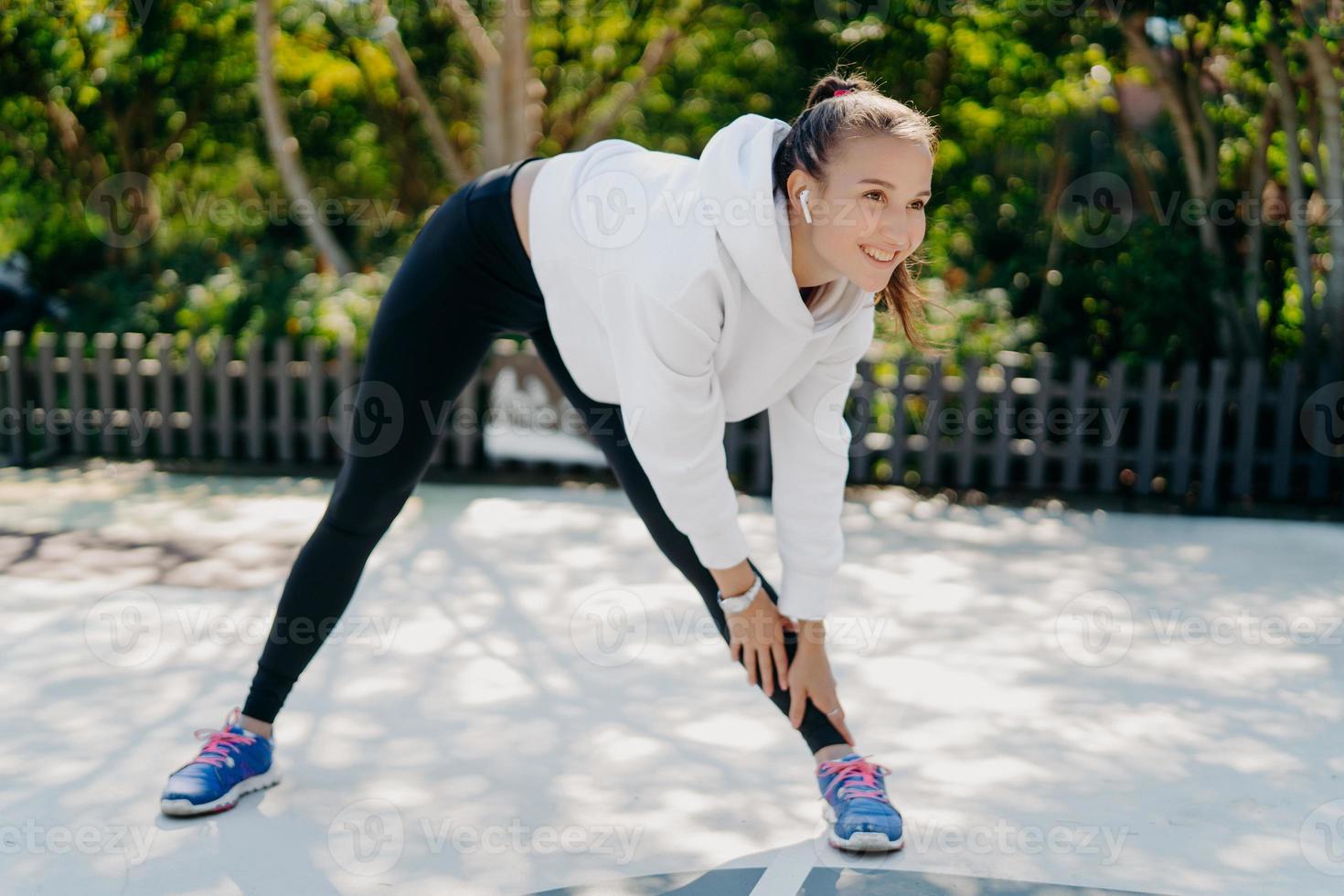 Photo of active pleased woman smiles gladfully concentrated into distance leans to leg dressed in sportswear trains outdoors being motivated listens music in wireless earphones does aerobic exercises