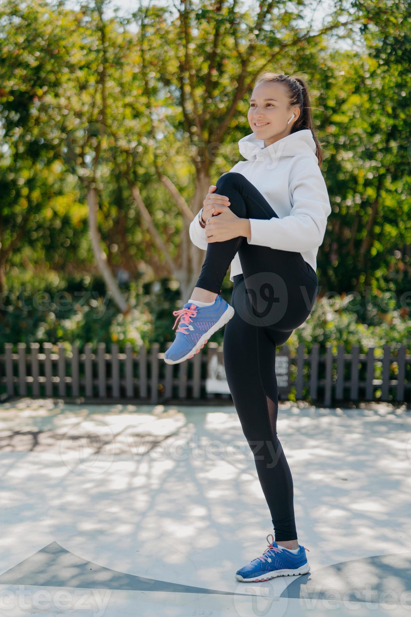 Full length shot of active healthy young woman warms up before jogging in  park keeps knee raised wears sweatshirt leggings and sneakers listens music  in earphones. Healthy lifestyle concept. 8240874 Stock Photo
