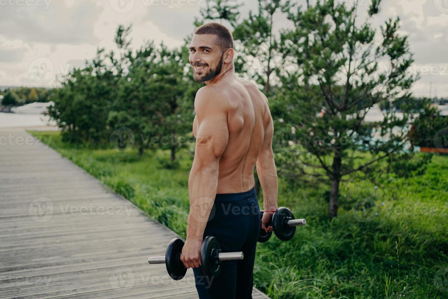 Muscular satisfied athlete bearded man gets energy during sport training, has strong arms, raises barbells, dressed in shorts, poses outdoor, enjoys regular workout in nature, builds healthy body. photo