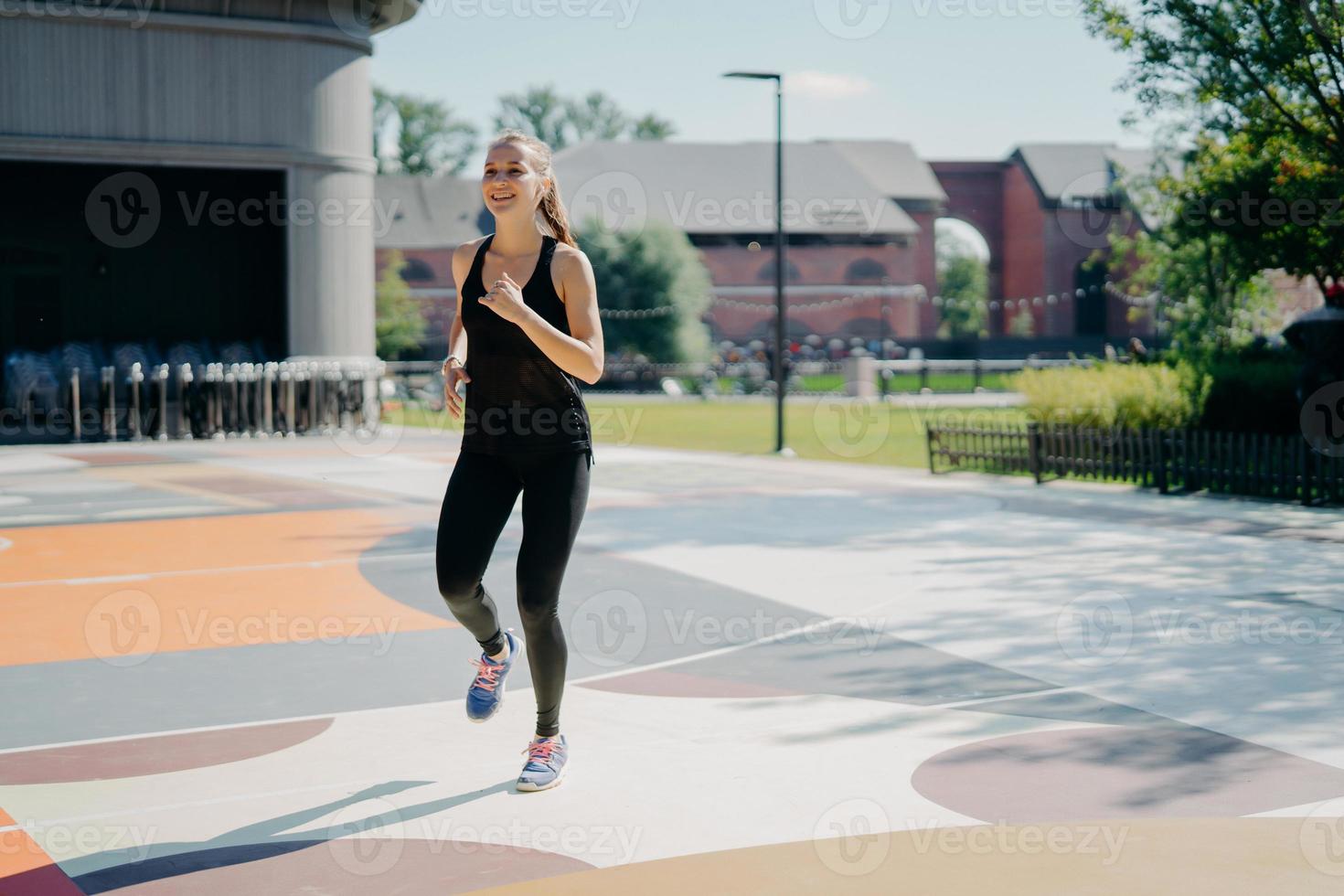 Cheerful athletic woman runs actively on stadium dressed in black sportclothes enjoys physical activities outdoors during summer day being full of energy. People sport and motivation concept photo