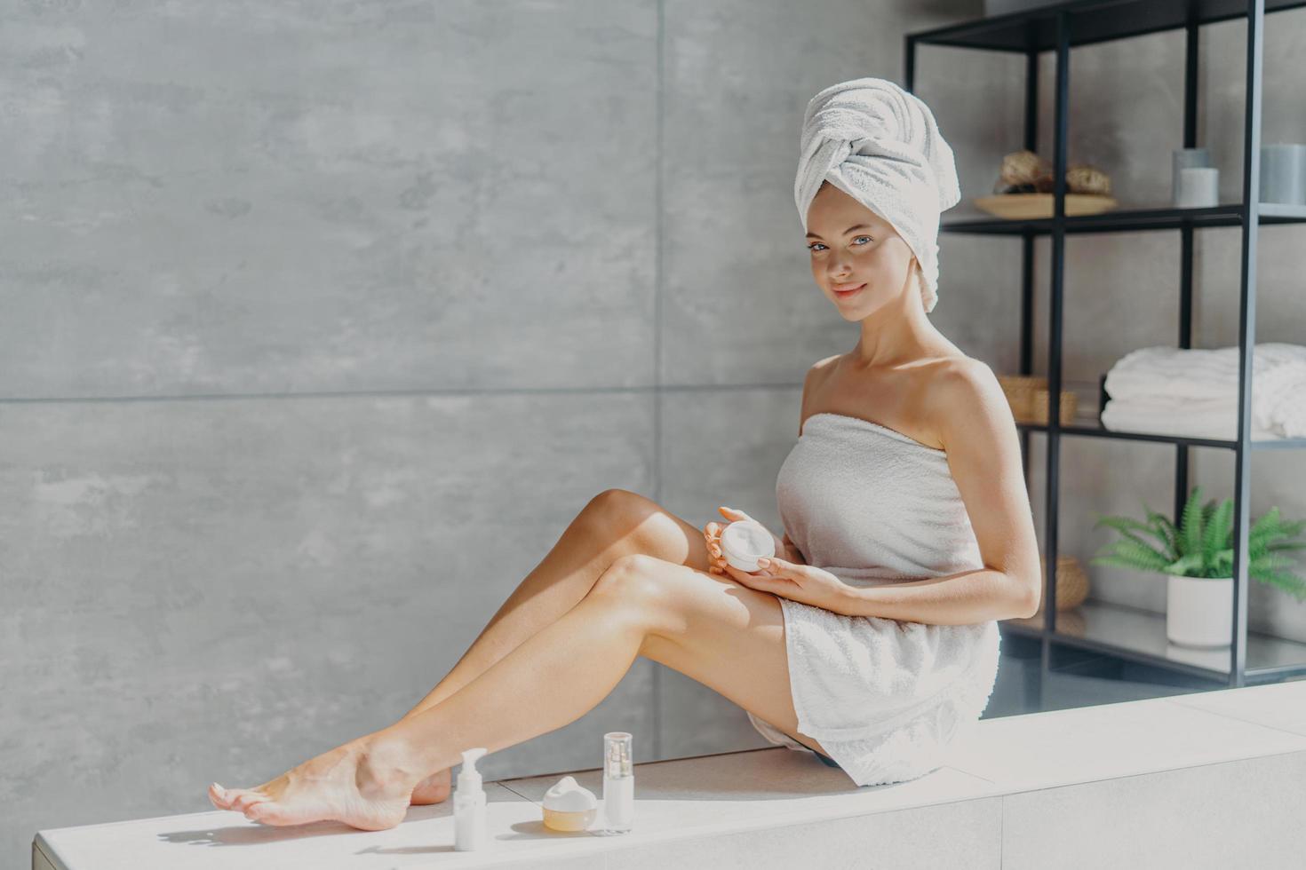 Photo of young female model enjoys skin care treatment, applies cream on legs, has healthy smooth glowing skin, well groomed body, poses wrapped in towel in bathroom, uses eco organic cosmetic