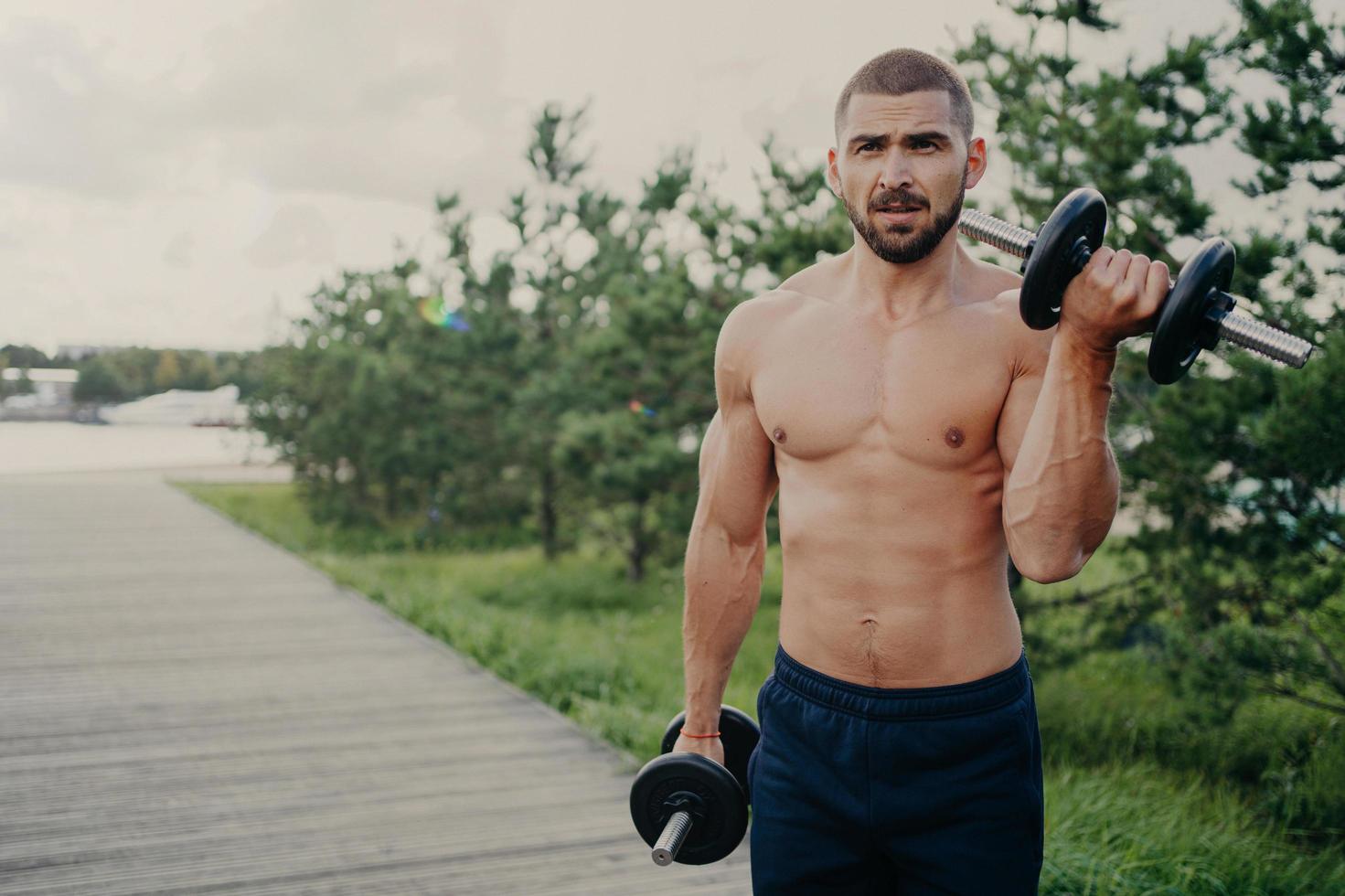 el culturista musculoso fuerte se prepara para la competencia atlética, levanta pesas pesadas, entrena al aire libre, demuestra su éxito deportivo, tiene el torso desnudo y el cuerpo perfecto. concepto de estilo de vida saludable foto