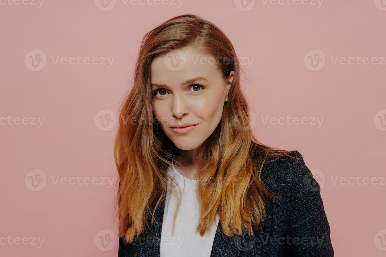 Attractive young woman with ginger hair in formal jacket posing against pink background photo