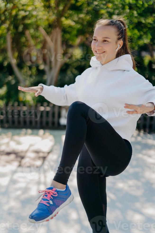 Fitness sport healthy lifestyle concept. Pleased sporty woman tries to balance on one leg keeps knee raised has interval training or cardio workout wears white sweatshirt leggings and sneakers photo