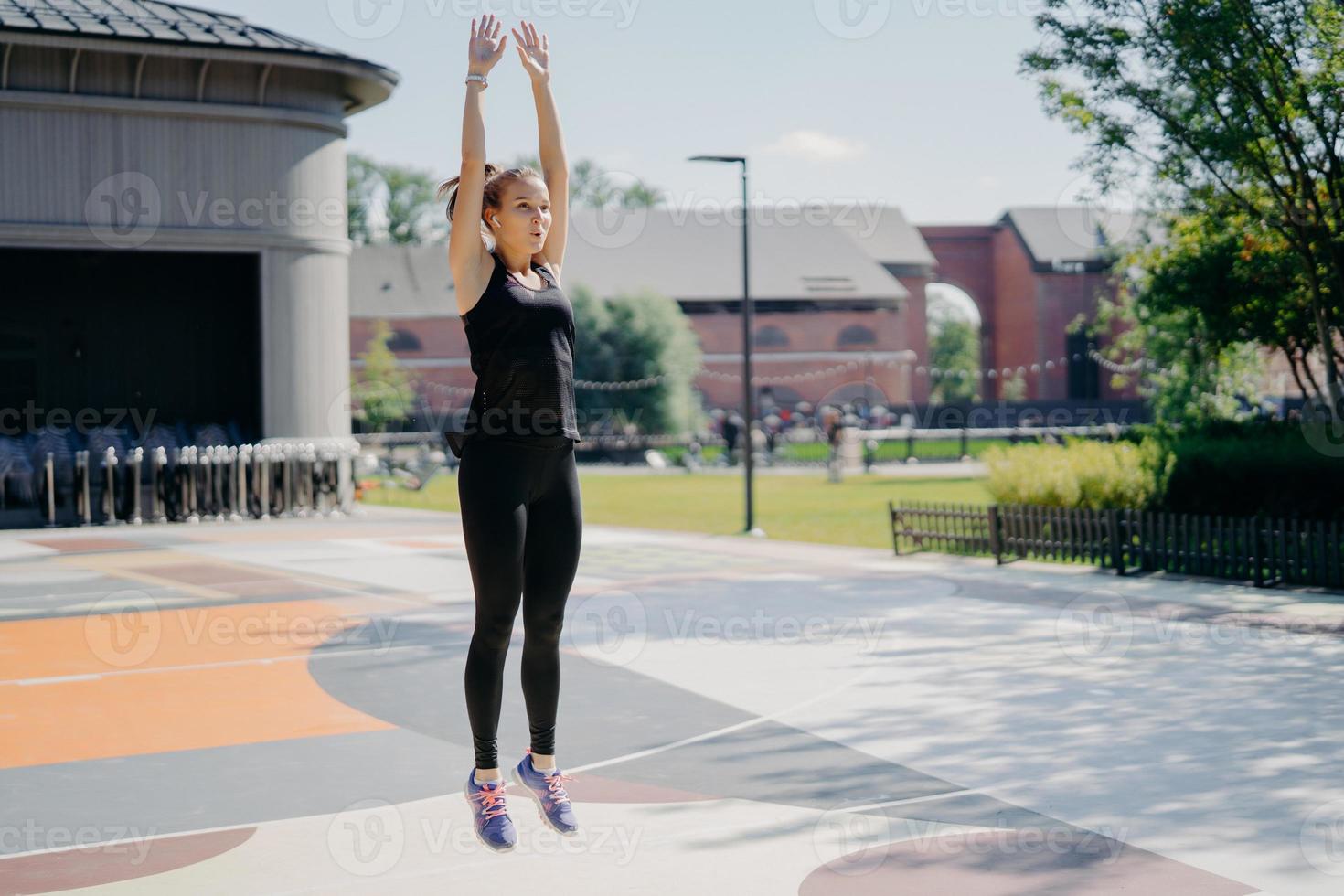 Full length shot of active young woman jumps high keeps arms raised up breathes deeply wears black t shirt leggings and sportshoes poses outdoors on stadium motivates you for going in for sport photo