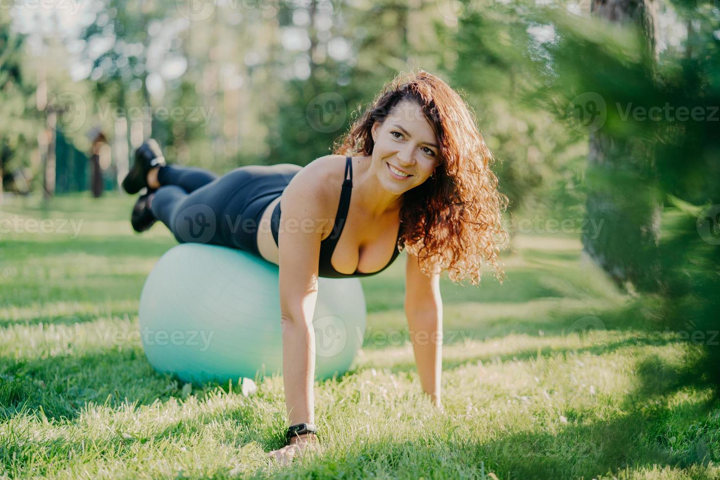 Happy active sportswoman does fitness exercises with gymnastic ball, dressed in active wear, has workout in open air, poses on green grass in forest or park, focused somewhere with pleasant smile photo