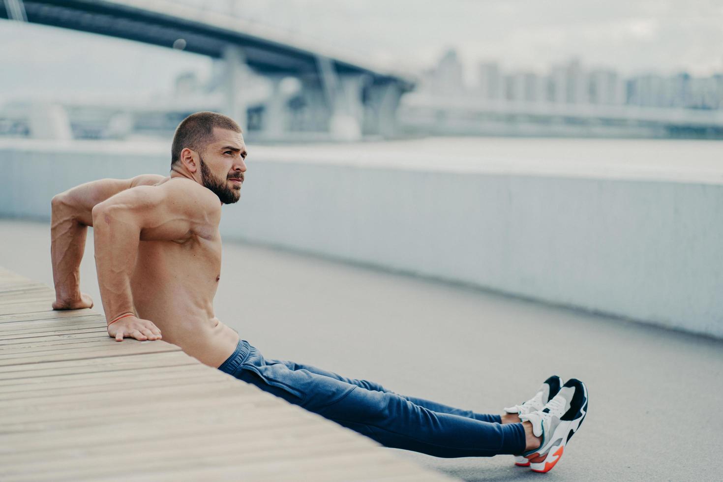 Handsome sportsman trains muscles and makes reverse push ups exercises warms up outdoors before run, concentrated aside, has muscular body, leads active healthy lifestyle, has bare muscular torso photo