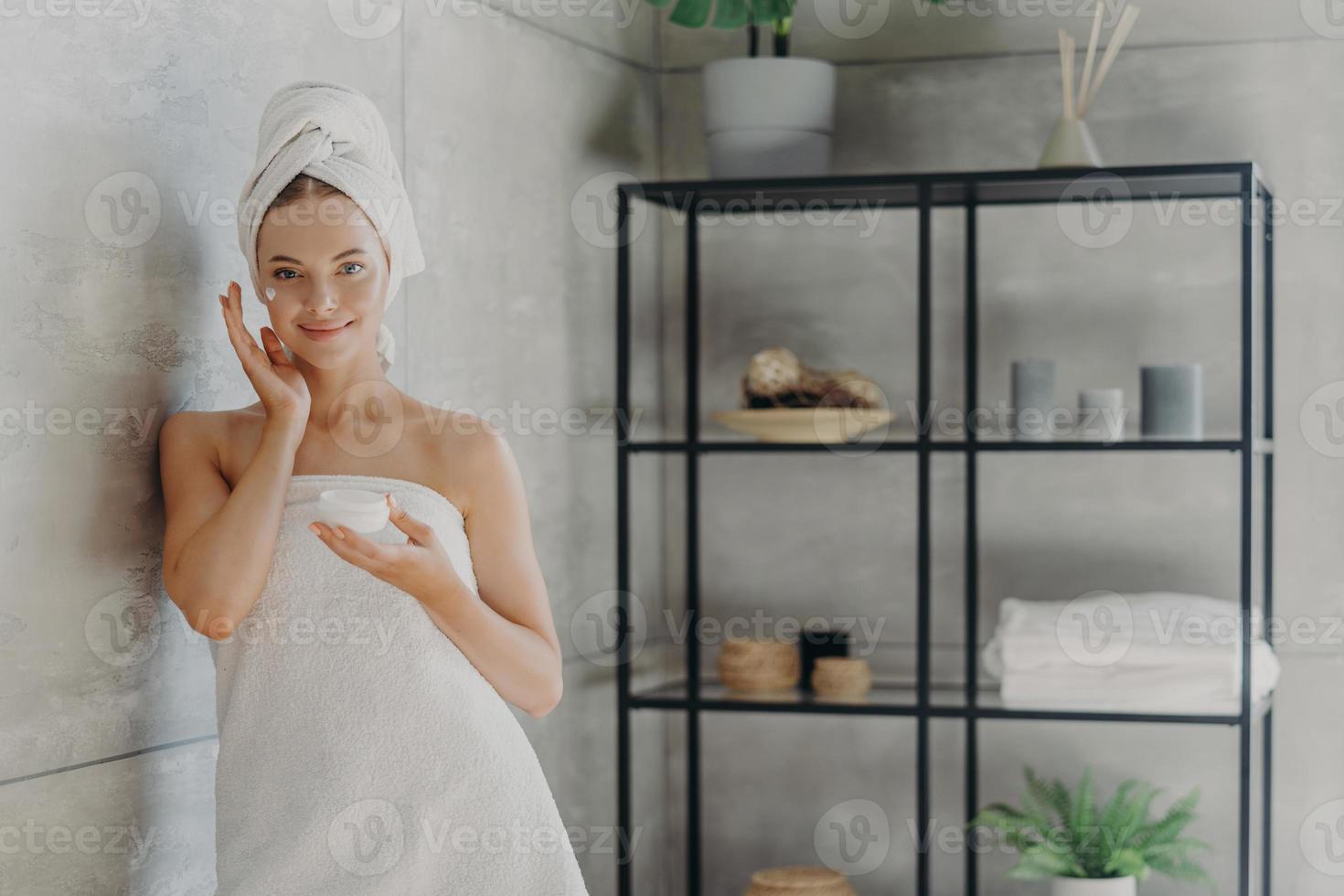 Horizontal shot of young healthy female model uses cream moisturizer, holds jar of cosmetic product, wrapped in towel after showering, stands near wall in cozy bathroom. Skin care procedures photo