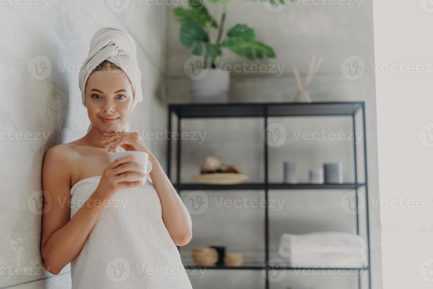 Pretty female model has perfect skin, touches jawline gently, drinks hot beverage, feels refreshed and relaxed, wrapped in bath towel, poses in bathroom. Natural beauty and skin care concept photo