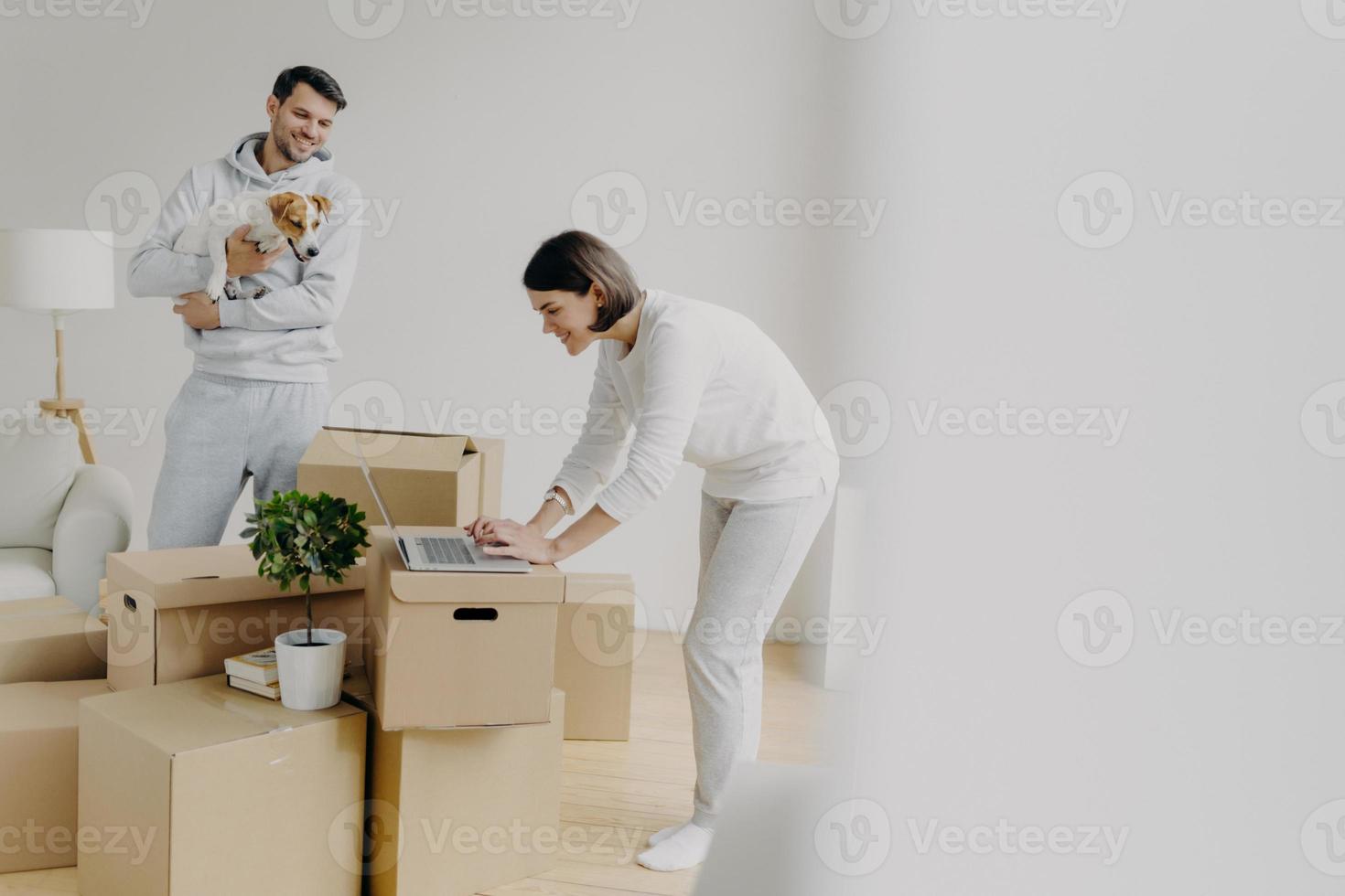 Indoor shot of attractive woman leans to laptop computer, focused into screen, surfes internet, searches ideas for decoration, man stands near, plays with favourite pet, move into new house. photo