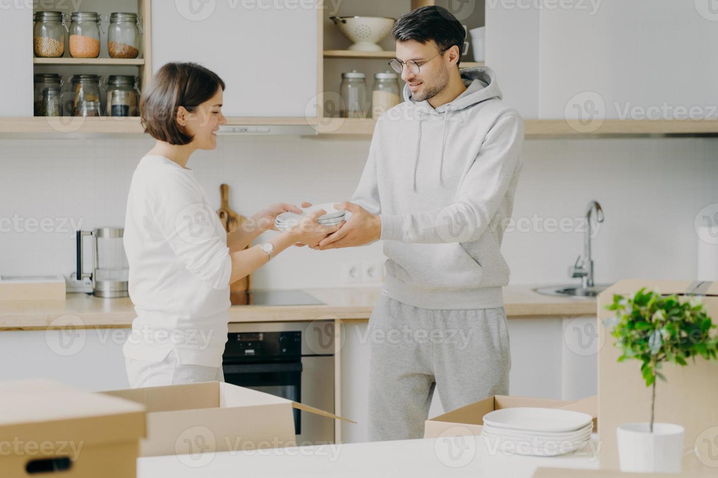 Horizontal shot of smiling couple unpack cardboard boxes in new kitchen, man passes plates to woman, placing dishes in cupboard, surrounded with moving packages in own bought or rented apartment photo