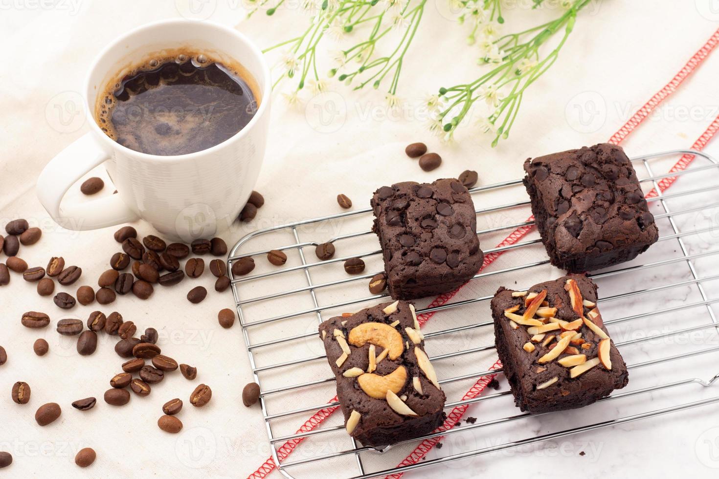 4 square dark brownies with chocolate chips, almonds and nuts on a baking rack, 1 white cup of coffee and coffee beans placed on a white cloth. photo