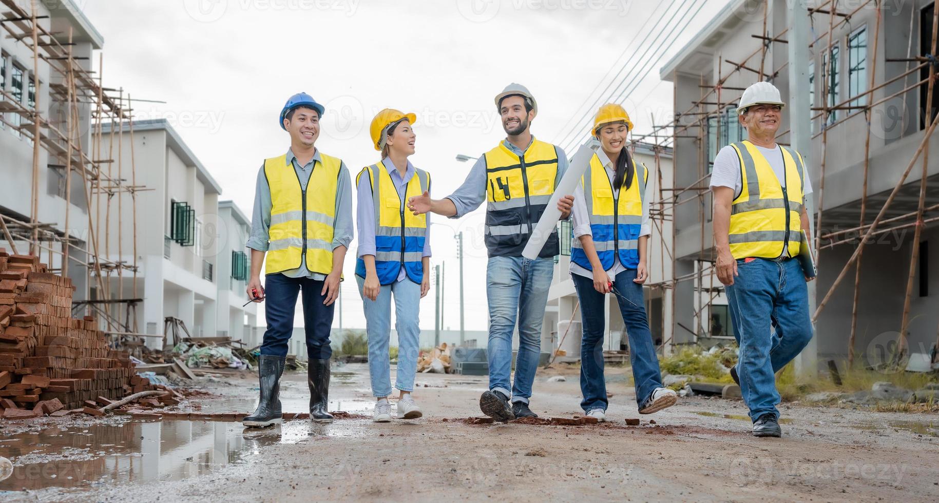 Team of engineers and workers wear safety clothing are walking through wet street in residential housing construction site. Engineering teamwork talking, discussing building project development. photo