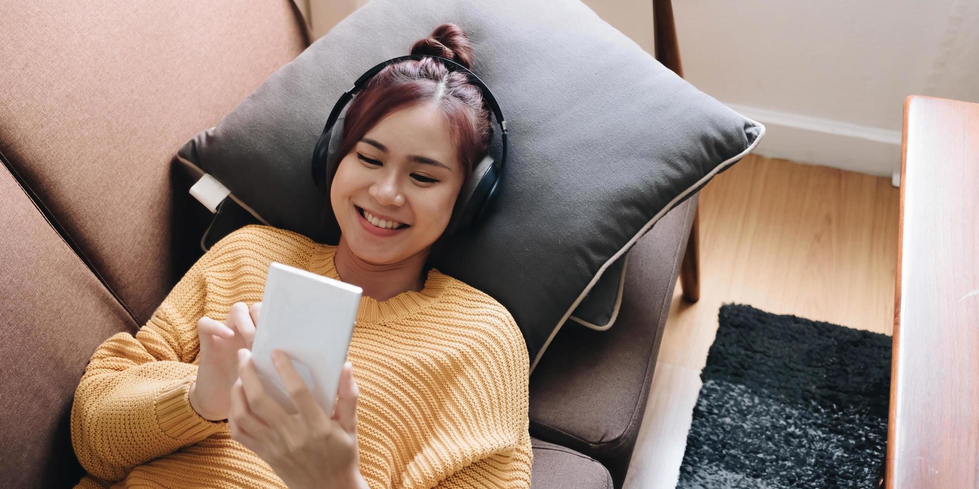 Lazy young woman spend weekend at home lying down on couch wearing headphones looking at smart phone screen listening music online. photo