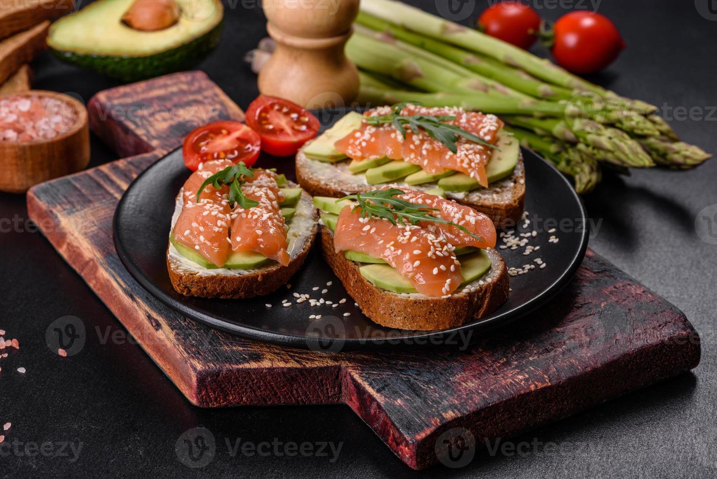 Toast sandwich with butter, avocado and salmon, decorated with arugula and sesame seeds, on a black stone background photo