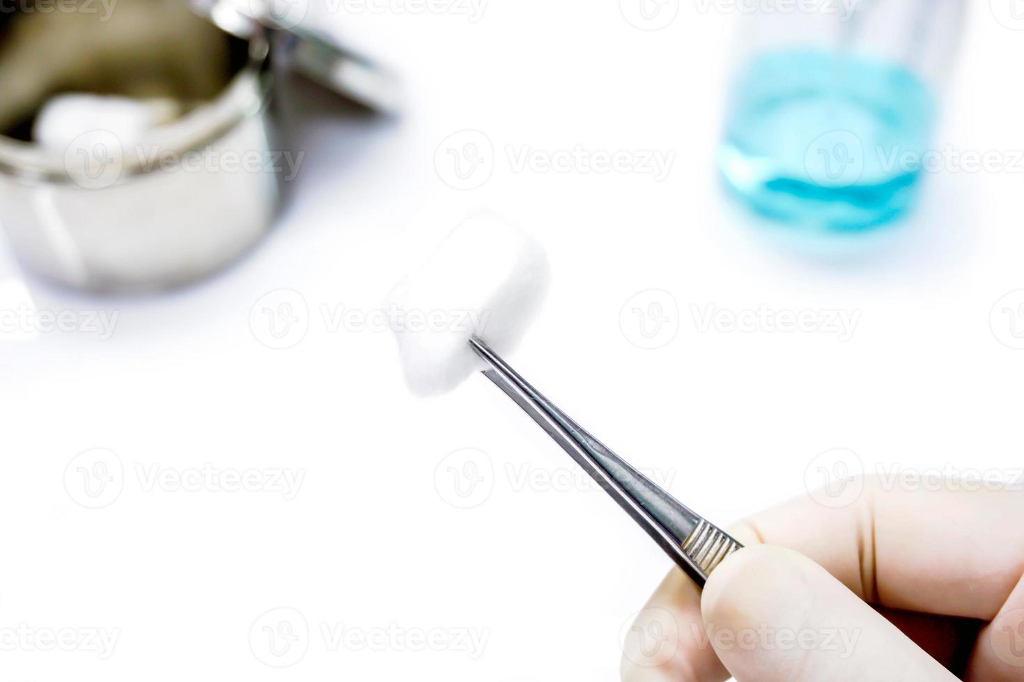 Doctor's hand  in the medical latex glove holding surgical forceps tong a cotton with blurry a cotton container and alcohol bottle on white background. photo