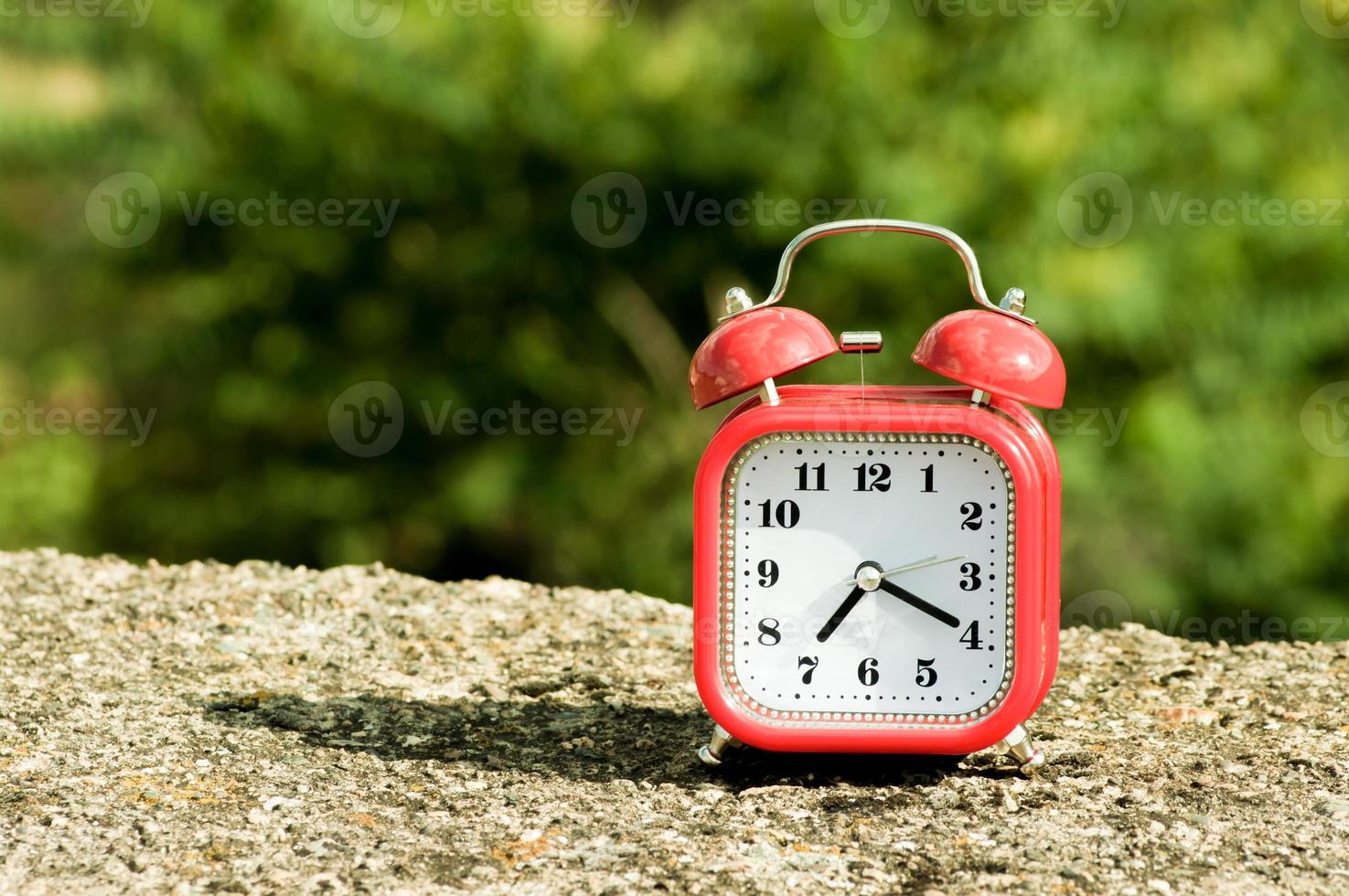 Red retro style alarm clock on a concrete surface with blurry trees on the background. photo