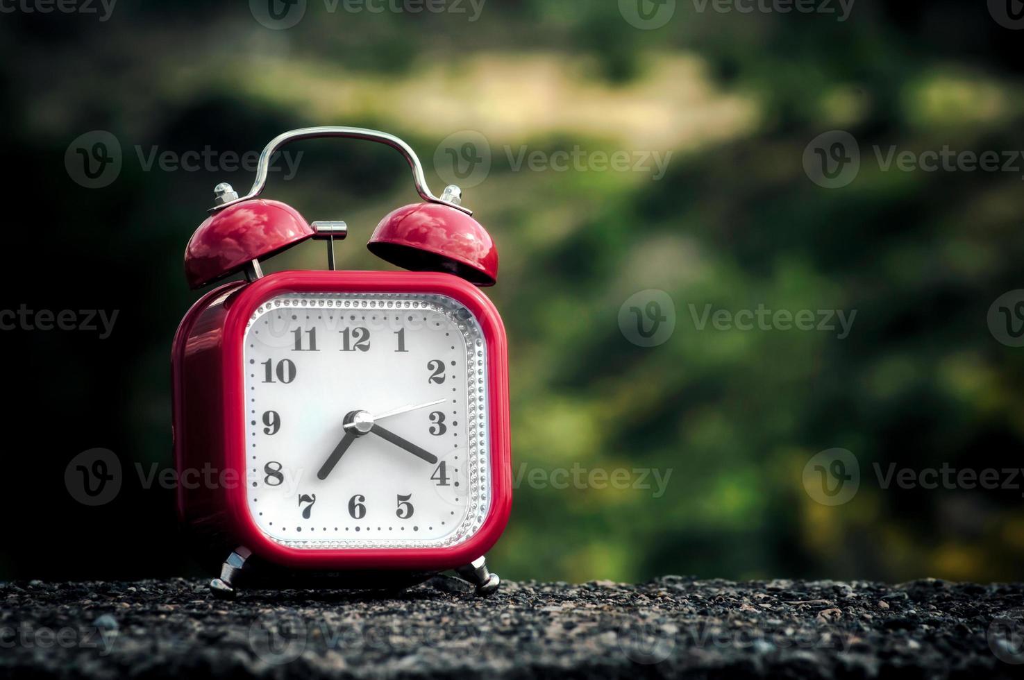 Red retro style alarm clock on a concrete surface with blurry trees on the background. photo