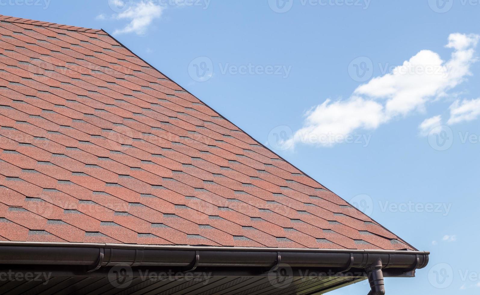 New roof with red shingles against the blue sky. High quality photo. Tiles on the roof of the house. Use to advertise roof fabrication and maintenance. Spotted texture. Affordable roofing. photo