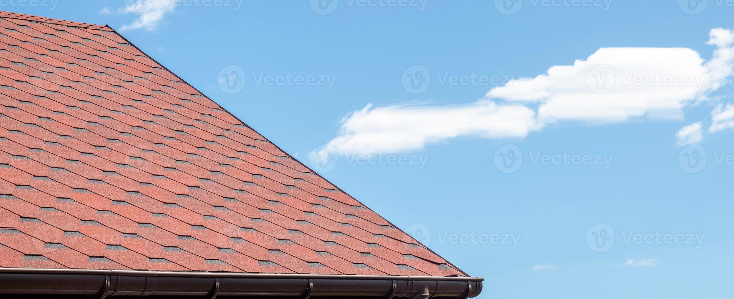 New roof with red shingles against the blue sky. Tiles on the roof of the house. Use to advertise roof fabrication and maintenance. Spotted texture. Affordable roofing. Banner with copy space. photo