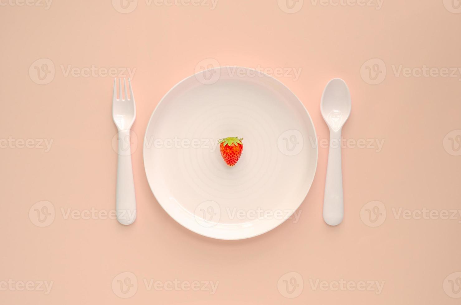 A strawberry fruit on white plate with fork and spoon on pink background. Minimal creative healthy diet food concept. photo