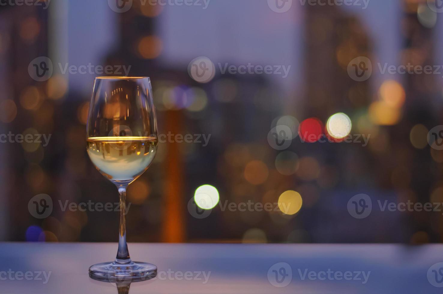A glass of white wine on table of rooftop bar with colorful bokeh of city light. photo