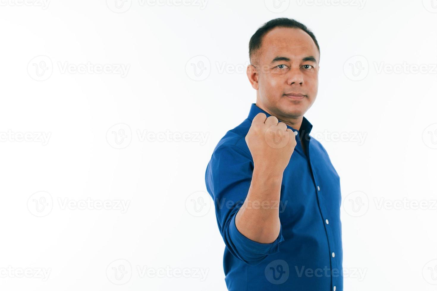 A self-confident middle age senior Asian man in a blue business casual shirt standing and showing the strong fist punch with copy space. Selective focus on hand. photo