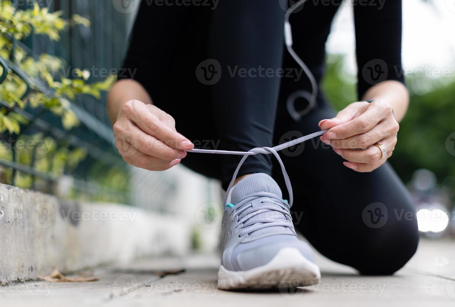 Crop unrecognizable woman runner wearing hygiene protective face mask sitting and tieing shoe rope during workout running in city park. Concept of healthcare in time of coronavirus outbreak photo