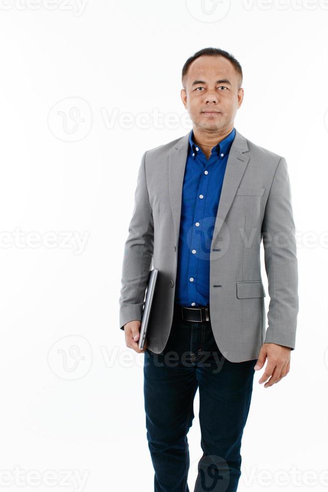 A self-confident middle age senior Asian man in a blue business casual shirt with a grey jacket standing and holding the notebook laptop computer and walking photo