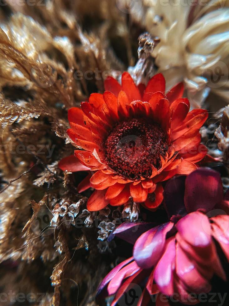 Red beautiful artificial flower on textured blurred background with dried plants, vertical photo. Decorative design interior element, closeup, wallpaper photo