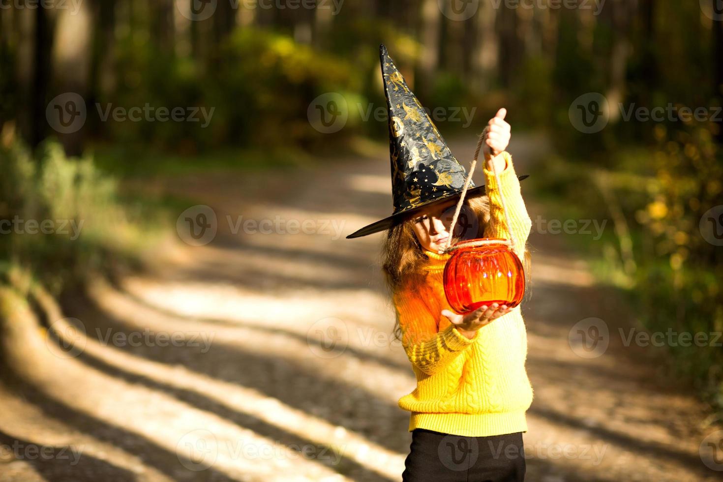 A girl in a witch's hat, in a yellow sweater and with a pumpkin a lantern and a bucket for sweets in autumn forest. Halloween Holiday. Portrait, close-up photo