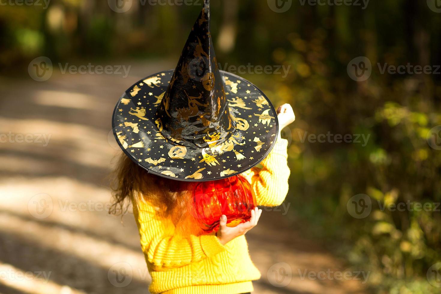 A girl in a witch's hat, in a yellow sweater and with a pumpkin a lantern and a bucket for sweets in autumn forest. Halloween Holiday. Portrait, close-up photo