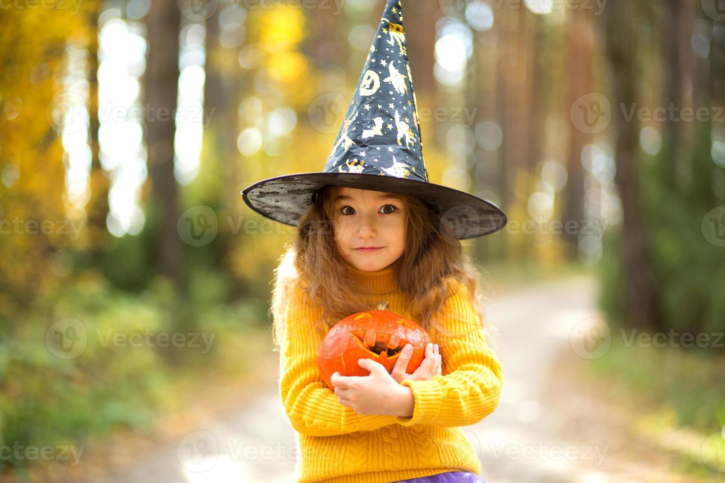 A girl in a witch's hat, in a yellow sweater and with a pumpkin with carved eyes and a mouth Jack o Lantern in a yellow autumn forest. Halloween Holiday photo