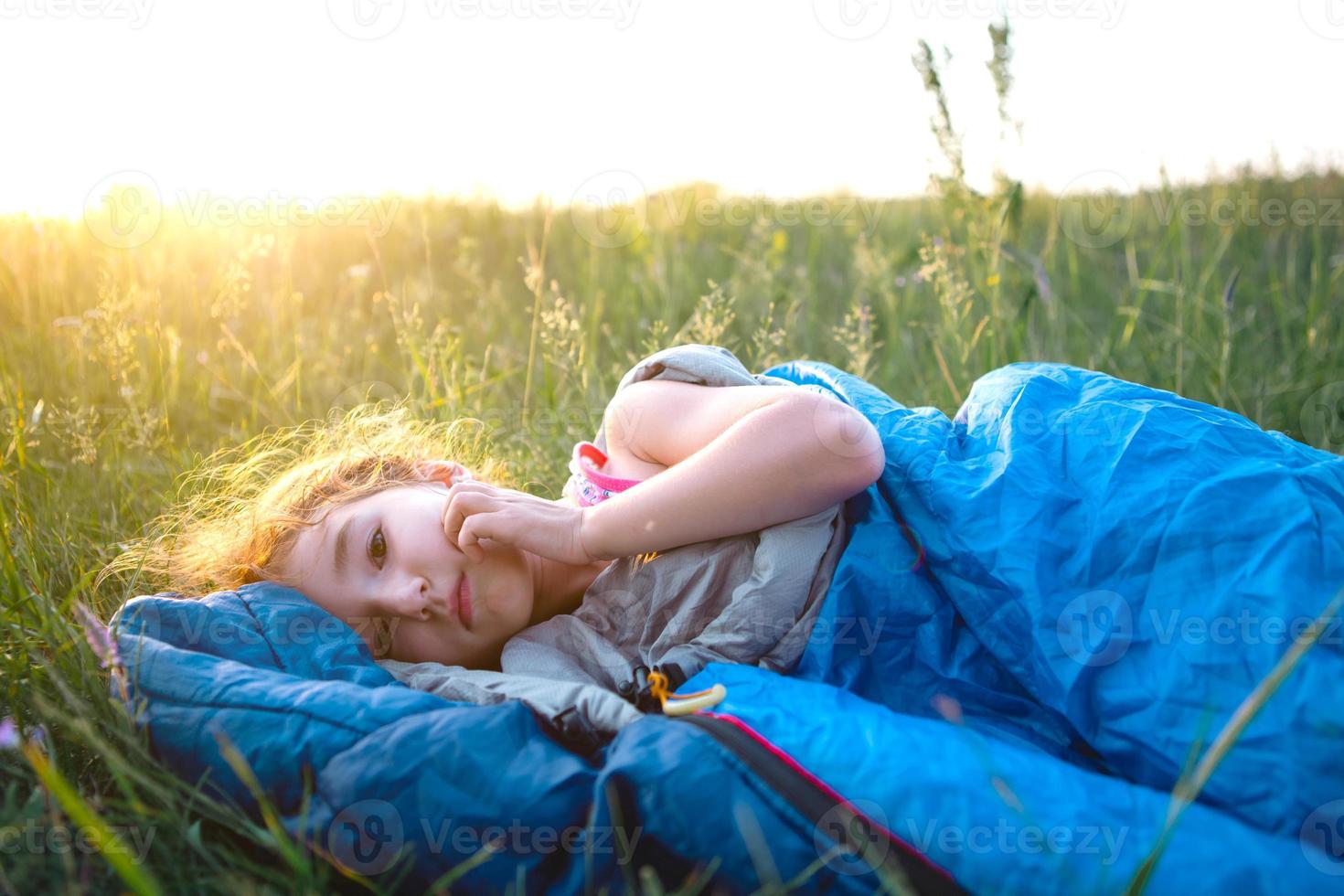 The girl is dissatisfied with scratching mosquito bites, child sleeps in a sleeping bag on the grass in a camping trip. Eco-friendly outdoor recreation, summer time. Sleep disturbance, repellent. photo