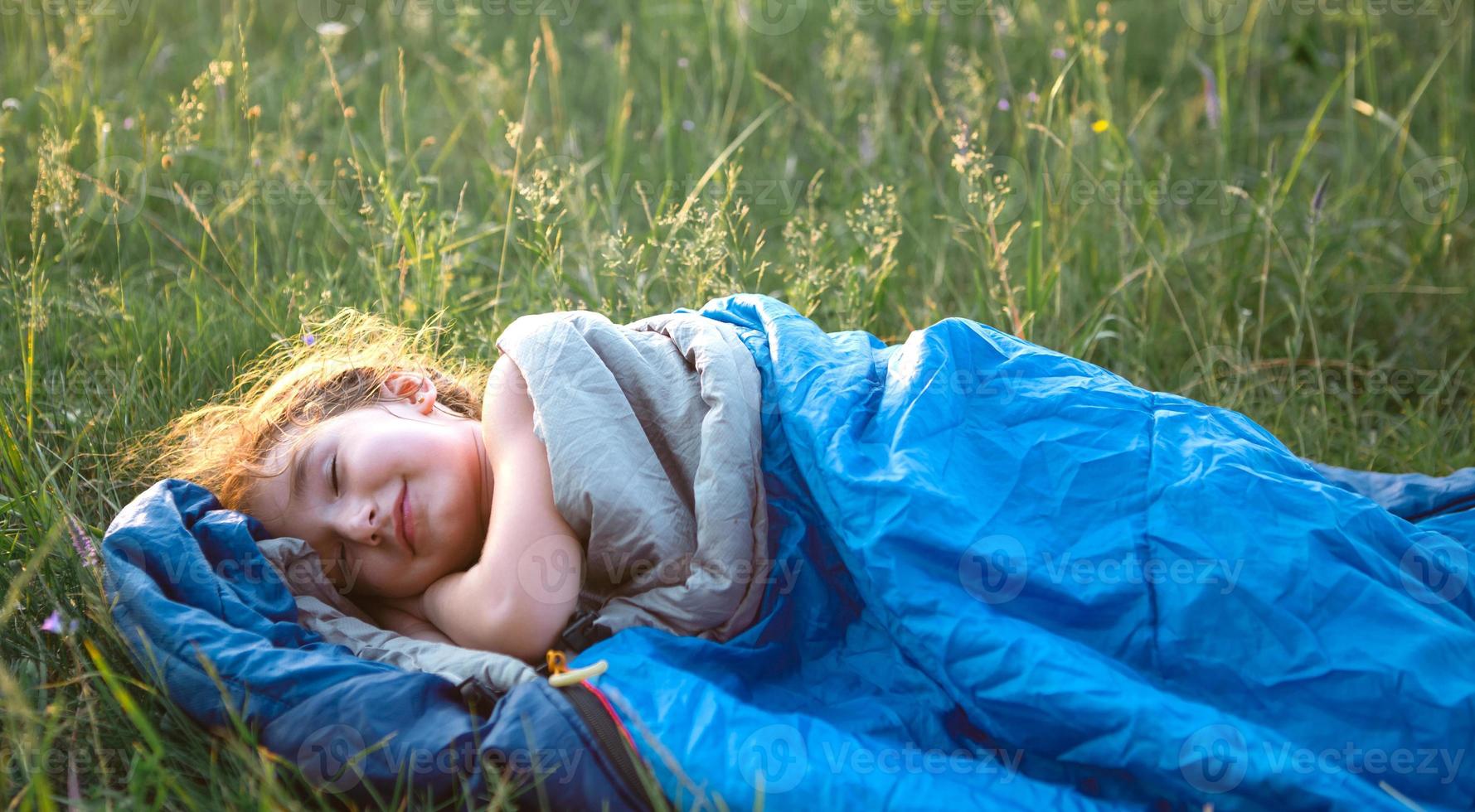 A child sleeps in a sleeping bag on the grass in a camping trip - eco-friendly outdoor recreation, healthy lifestyle, summer time. Sweet and peaceful sleep. Mosquito bites, repellent. photo