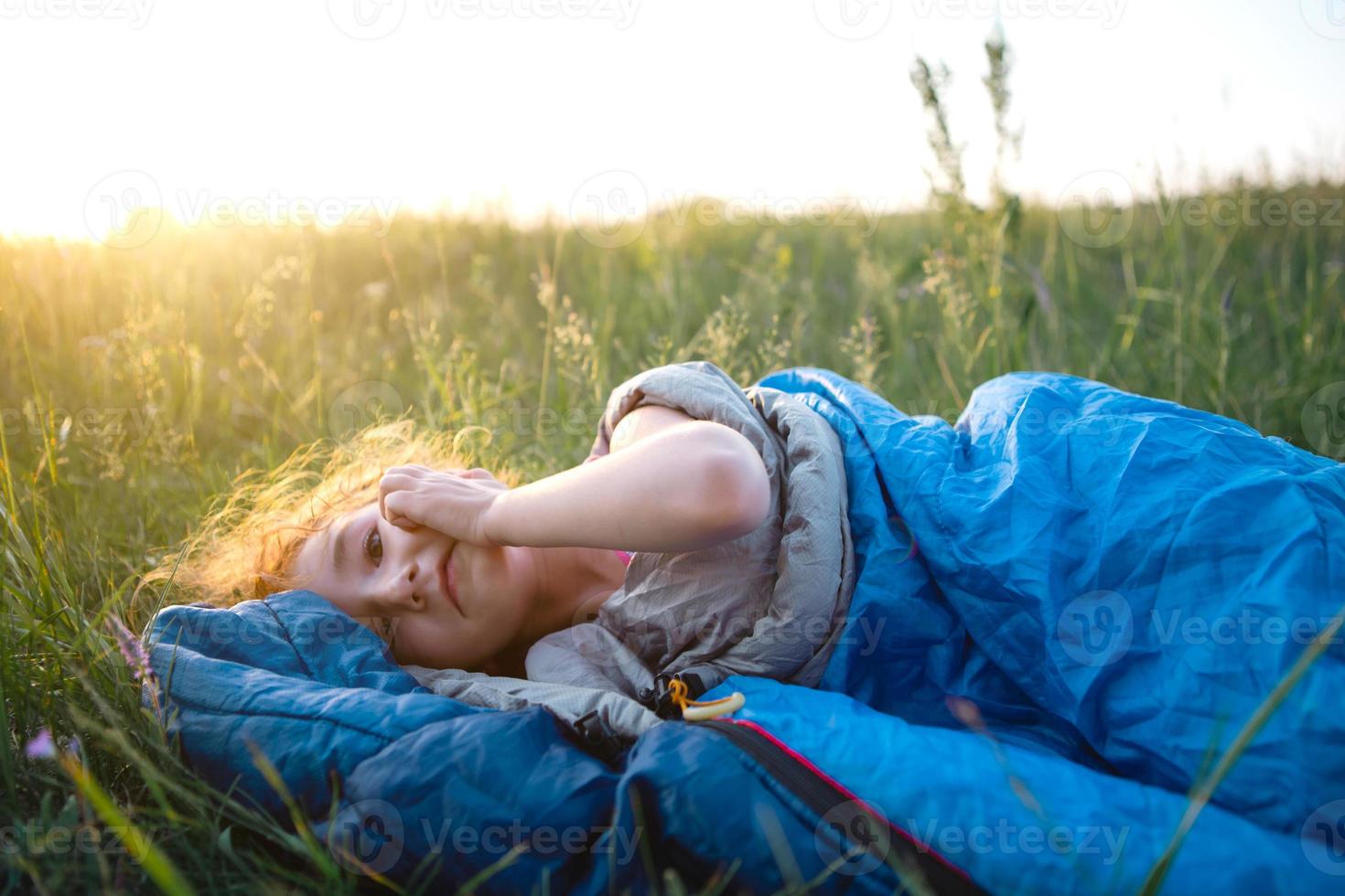 The girl is dissatisfied with scratching mosquito bites, child sleeps in a sleeping bag on the grass in a camping trip. Eco-friendly outdoor recreation, summer time. Sleep disturbance, repellent. photo