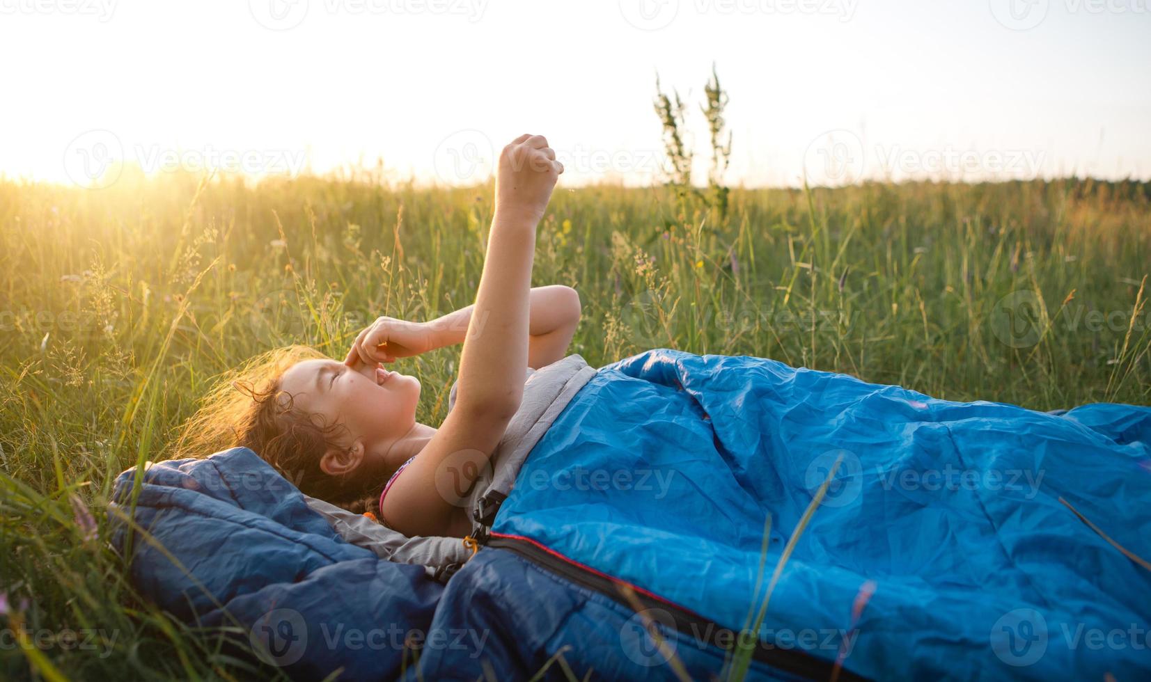 The girl is dissatisfied with scratching mosquito bites, child sleeps in a sleeping bag on the grass in a camping trip. Eco-friendly outdoor recreation, summer time. Sleep disturbance, repellent. photo