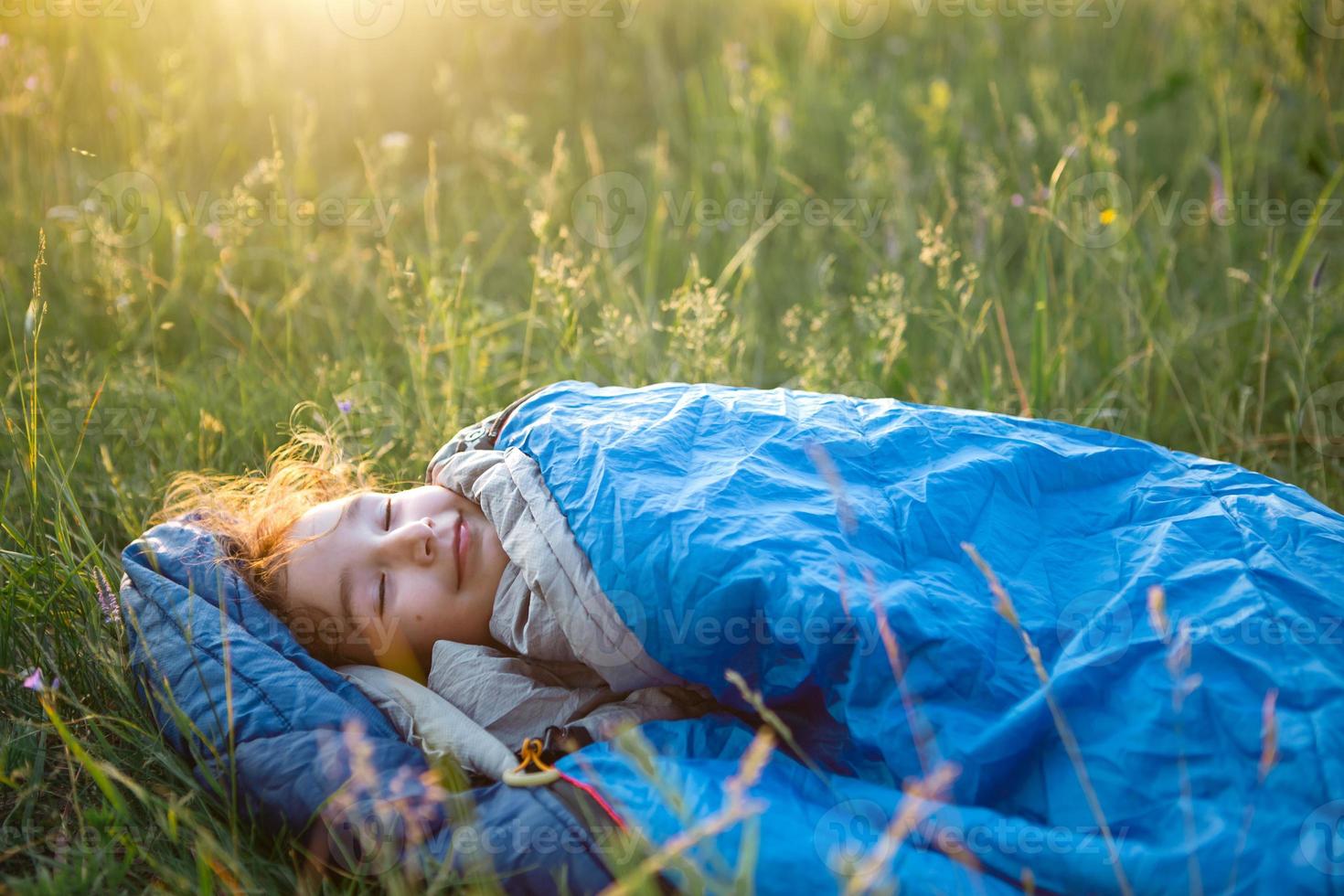 A child sleeps in a sleeping bag on the grass in a camping trip - eco-friendly outdoor recreation, healthy lifestyle, summer time. Sweet and peaceful sleep. Mosquito bites, repellent. photo