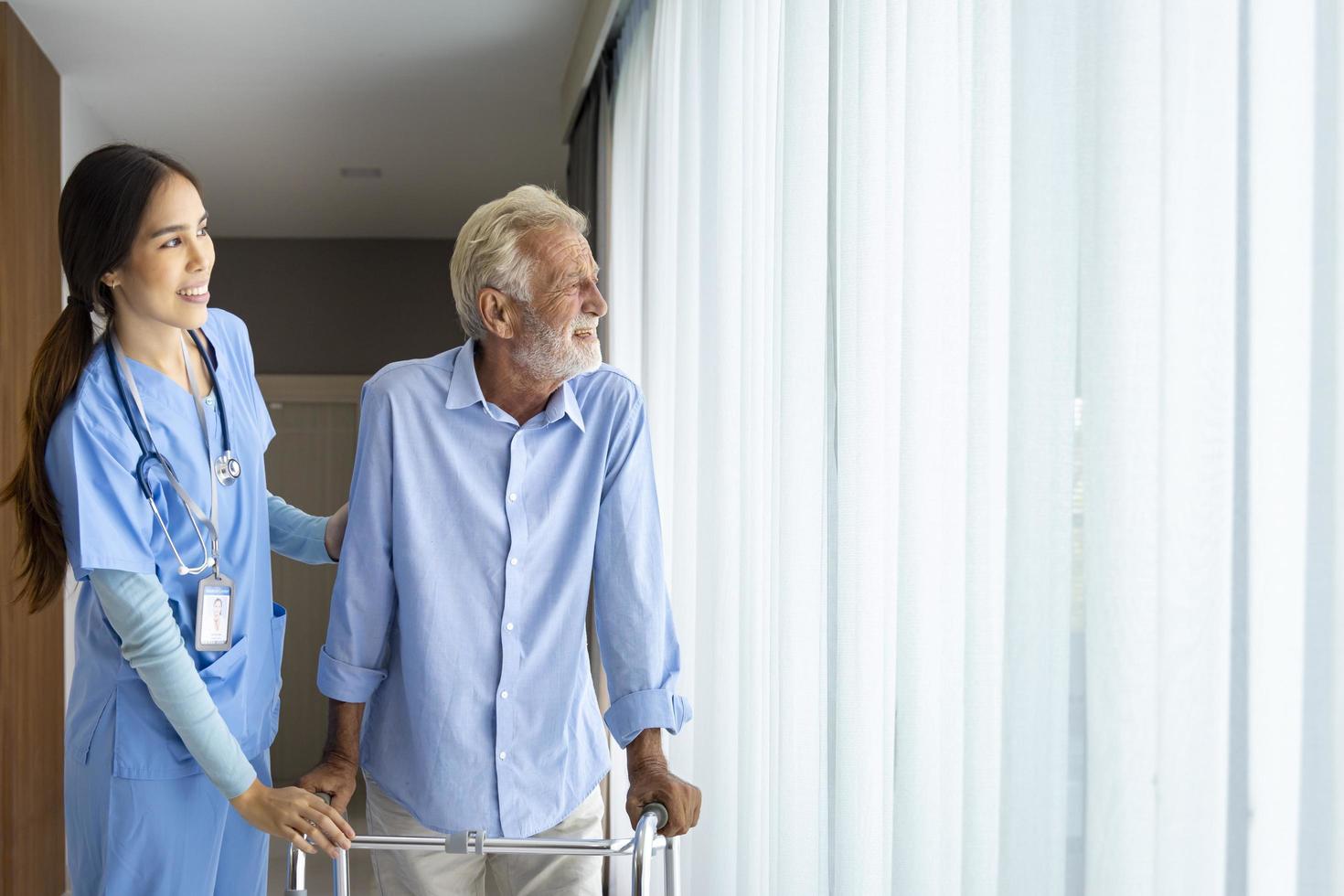 Hospice nurse is supporting Caucasian man to use walker while looking out the window in pension retirement center for home care rehabilitation and post treatment recovery process photo