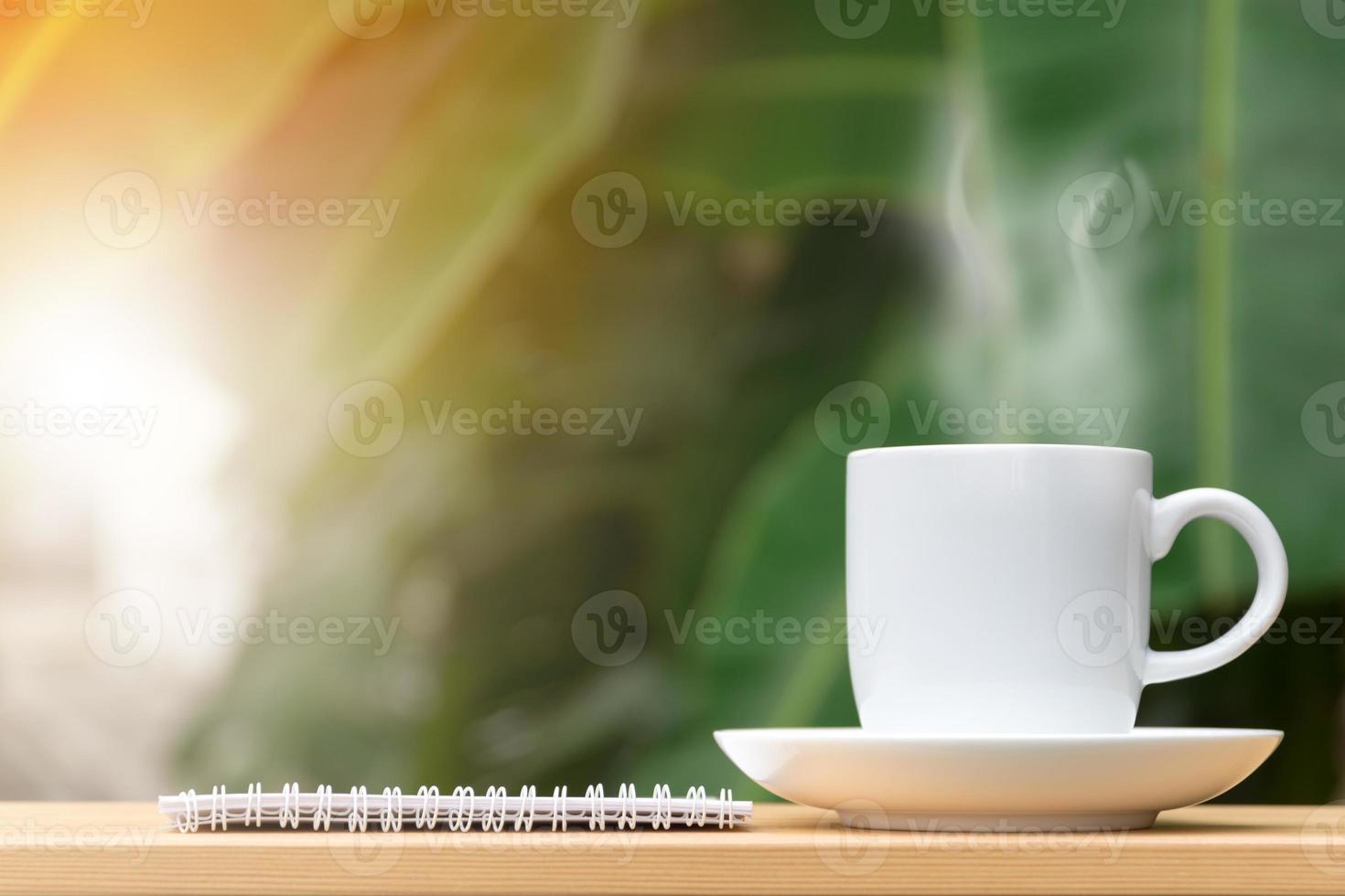 White coffee cup with stream and notebook on wooden table garden in background. photo