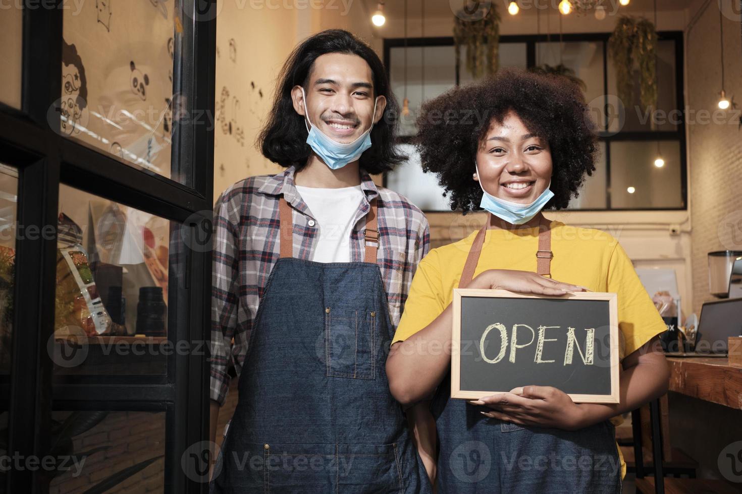Two young startup barista partners stand at casual cafe door, write and show open sign before taking off face masks, happy and cheerful smiles with coffee shop service jobs after COVID-19 lifestyle. photo